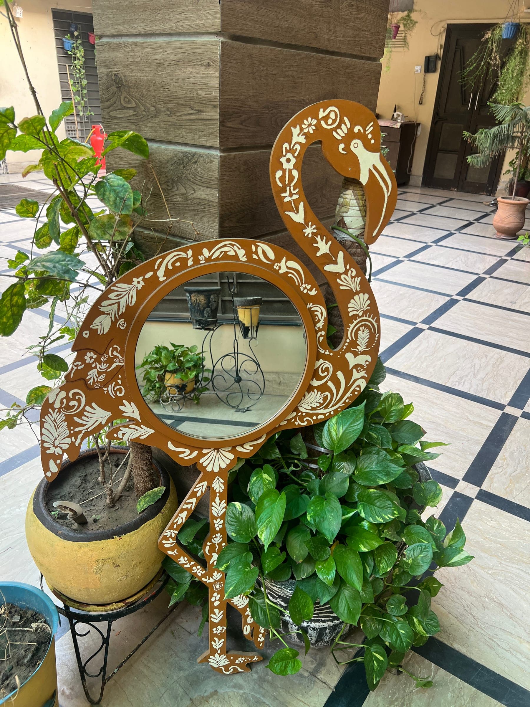 The Flamingo Mirror beside potted plants, its round mirror reflecting the courtyard