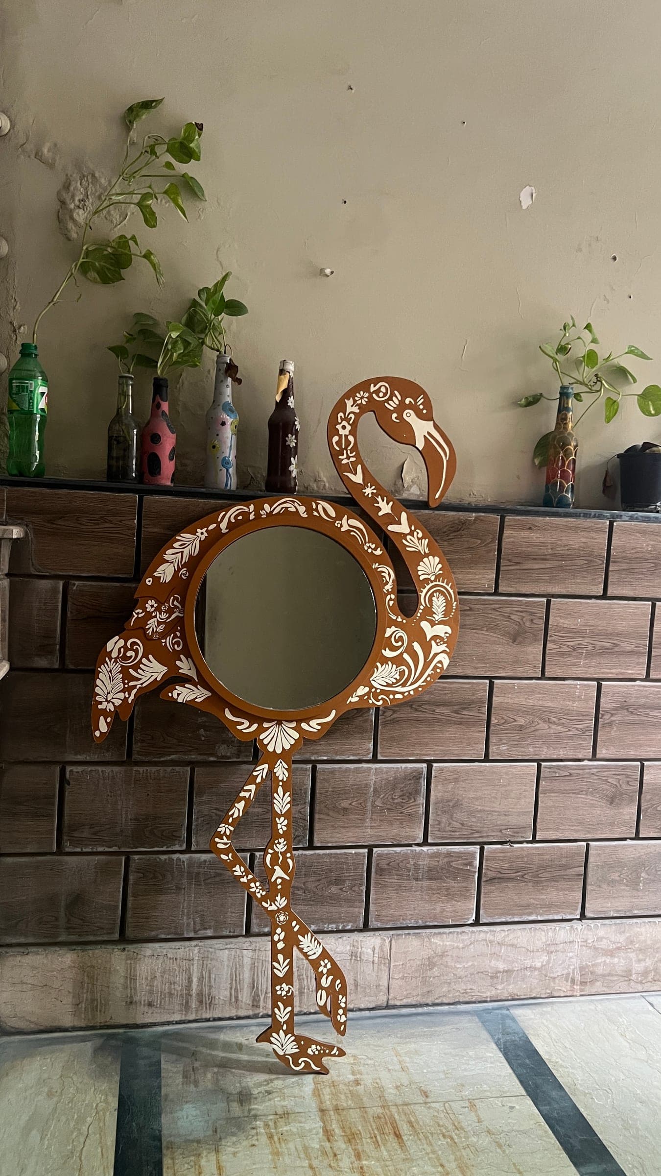 The Flamingo Mirror on a brick wall below a shelf of painted bottles