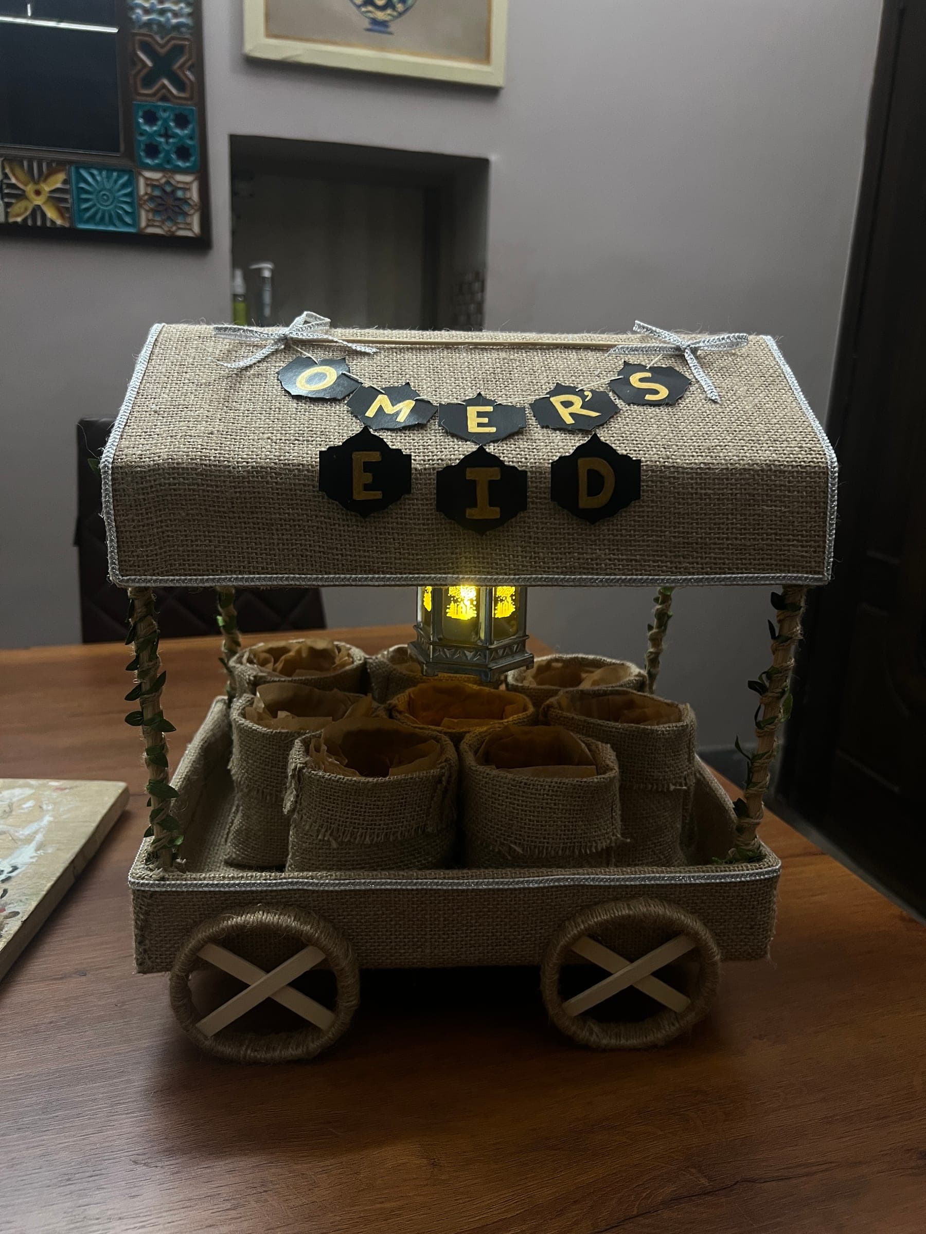Eid gift cart with a jute canopy and wooden wheels, filled with jute pouches, under a personalised Eid bunting
