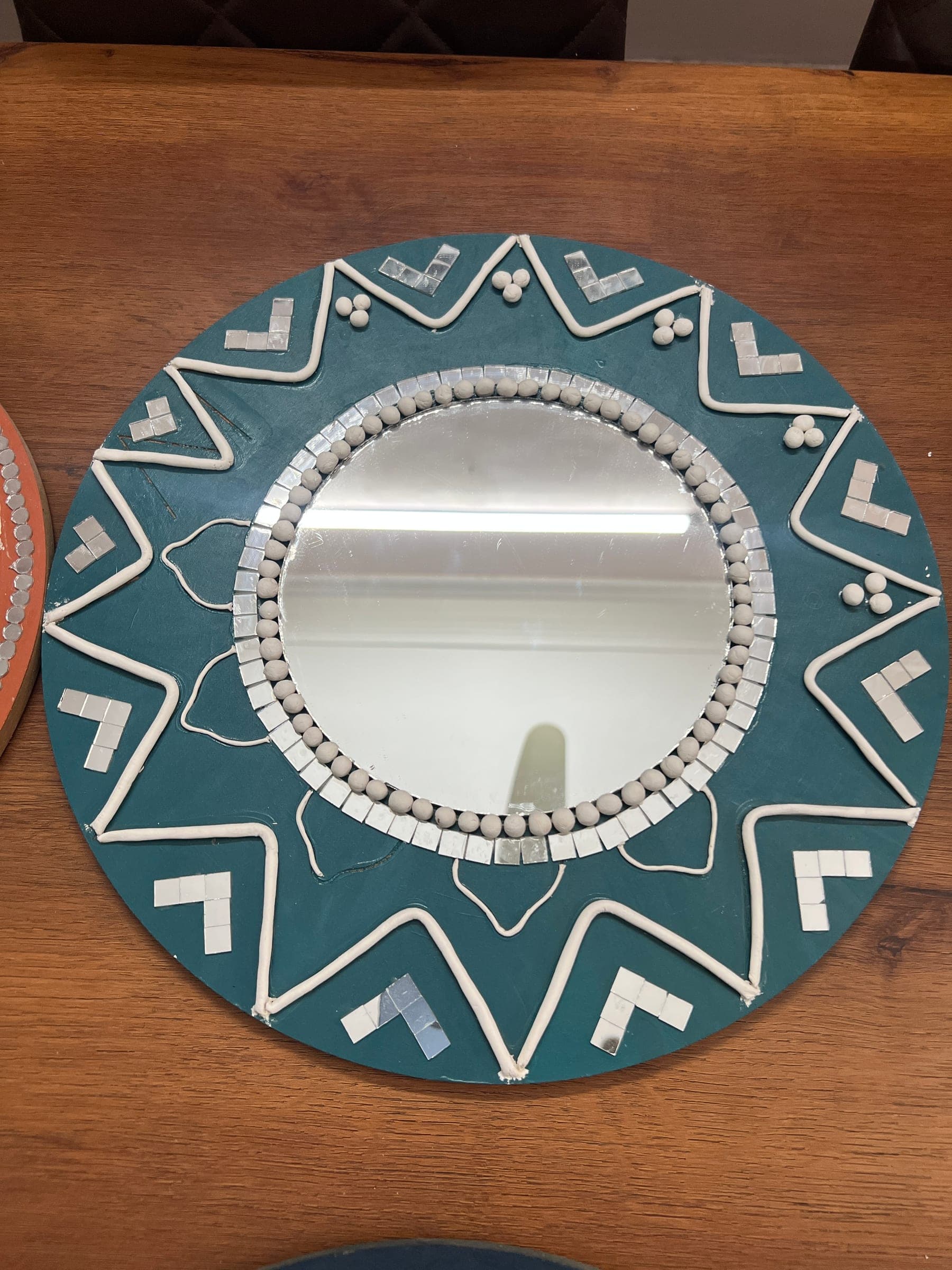 Teal Lippan mirror: a round mirror framed by white clay zigzags and mirror chips on a teal ground